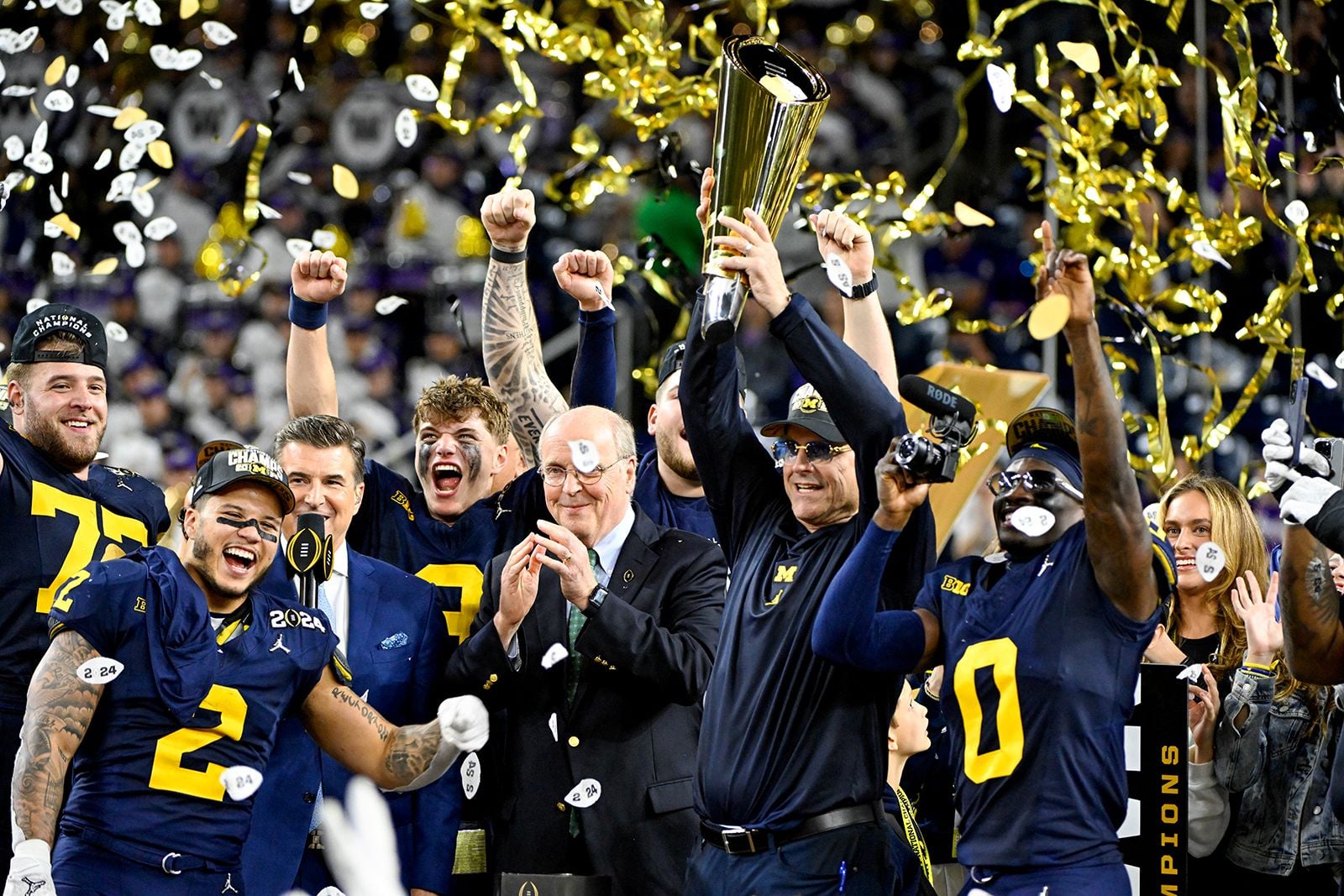 Football players and staff in navy and maize uniforms celebrate a championship victory, with a Jim Harbaugh holding a gold trophy amid confetti and cheering fans.