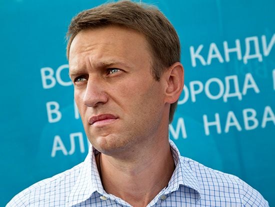 Aleksey Navalny in a checkered shirt looks to the side with a serious expression. A blue background with partially visible white text is behind him.