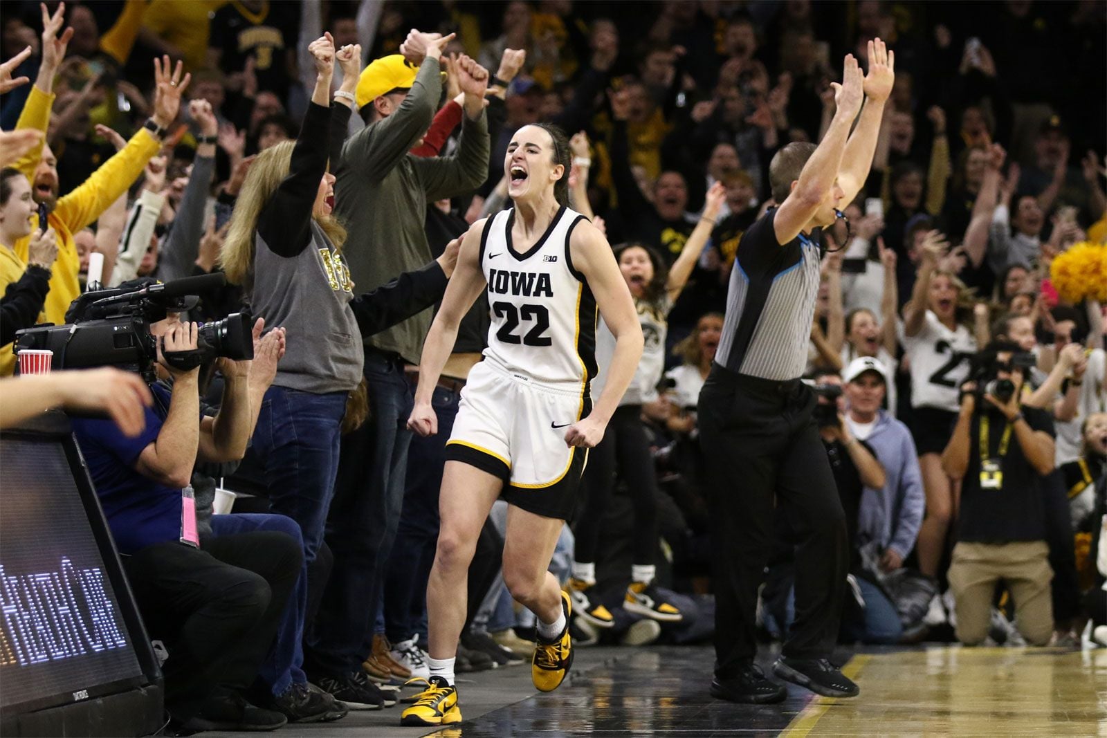 Caitlin Clark in an Iowa uniform (#22) celebrates on the court with clenched fists as fans and officials cheer in the background.