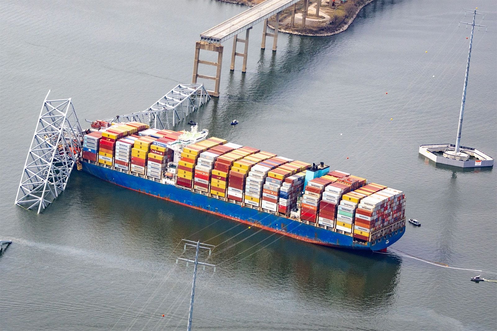 A cargo ship loaded with colorful shipping containers is lodged in a collapsed bridge structure over a body of water, with small boats nearby.