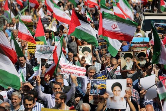 A crowd of protesters holding Iranian flags, Palestinian flags, and various signs, including portraits of leaders and anti-Israel slogans, in a public demonstration.