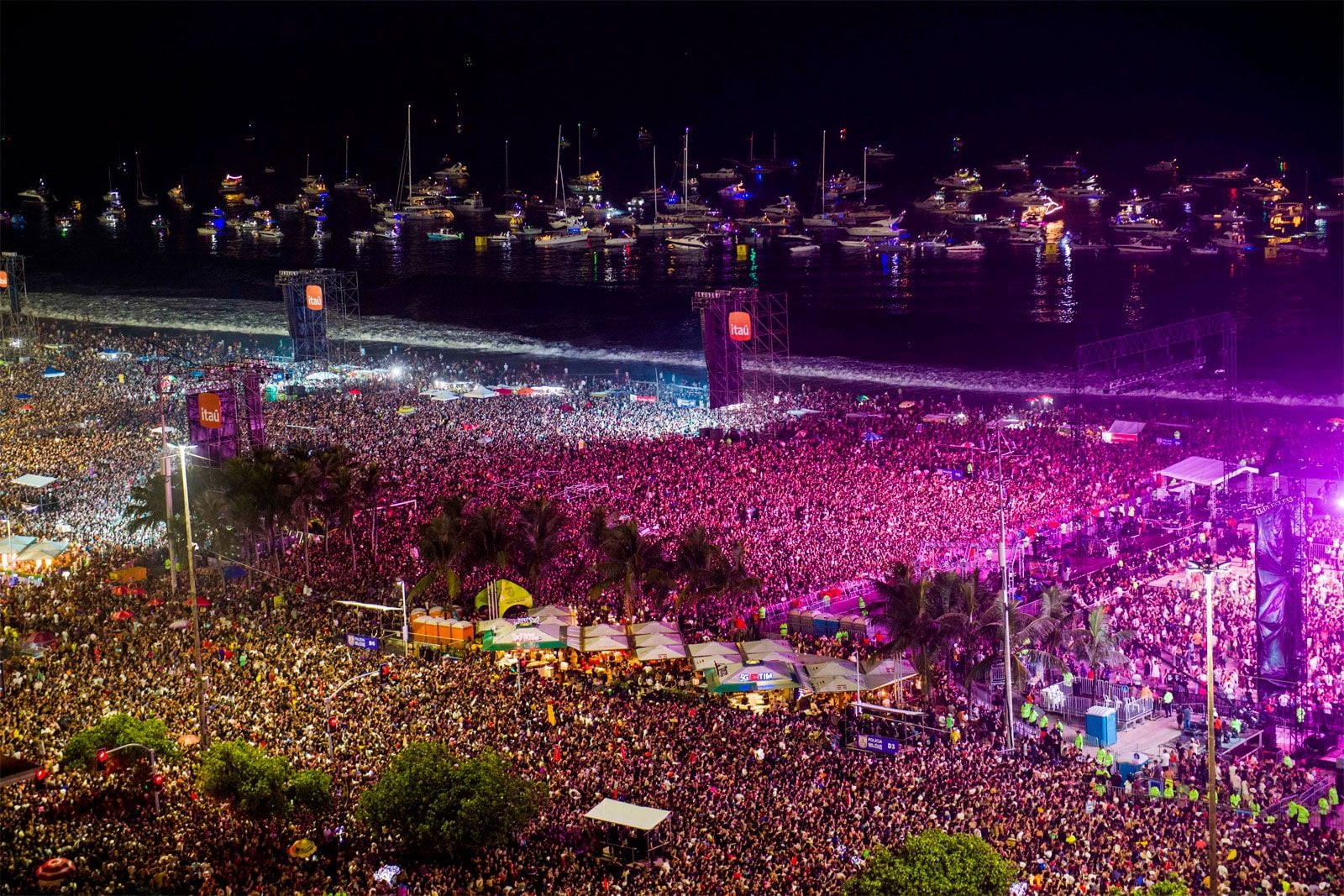 A massive nighttime crowd at a waterfront festival with colorful lights, stages, and boats visible on the water in the background.