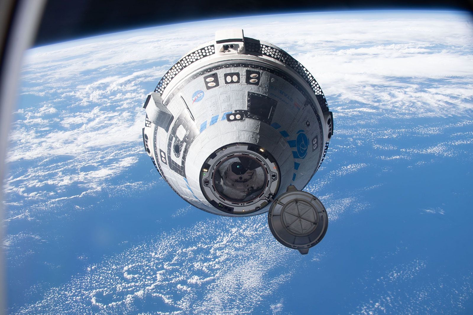 Boeing's Starliner with an open hatch floating above Earth's surface, showing clouds and the blue ocean below, viewed from another spacecraft window.