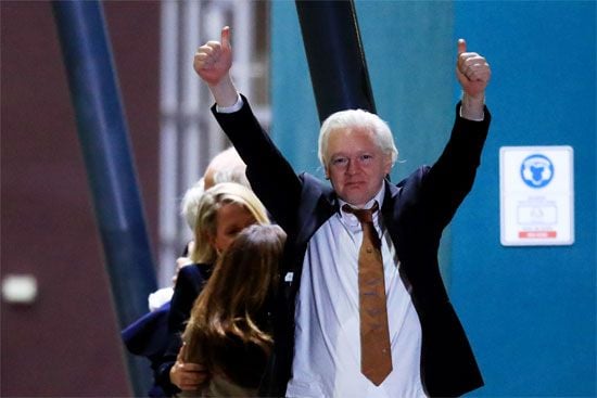 WikiLeaks founder Julian Assange, in a suit with a loosened tie, raises both thumbs up upon arriving at Canberra Airport on June 26, 2024, in Canberra, Australia, with others nearby and a building in the background.