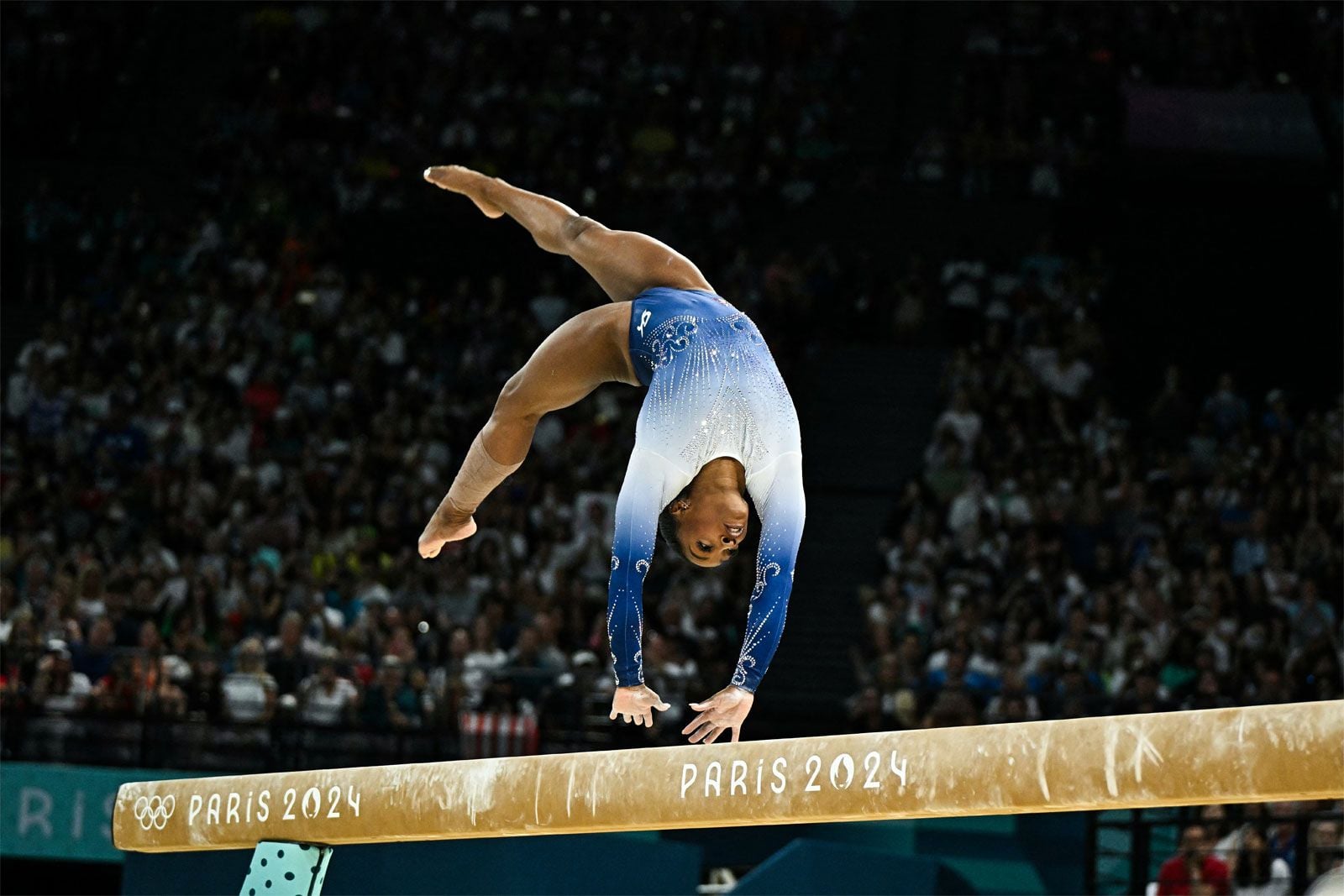 Simone Biles performs a handstand on the balance beam during the Paris 2024 Olympics, wearing a blue and white leotard, with a crowd in the background.