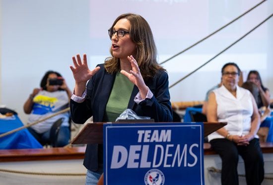 Sarah McBride speaks passionately at a podium with a "Team DELDEMS" sign. People sit in the background, some listening and others taking photos.