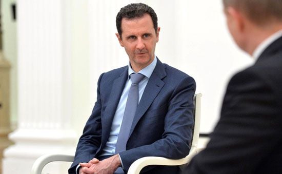 Bashar al-Assad wearing a navy suit, light blue shirt, and tie, sits on a white chair in conversation with another person whose back is facing the camera.